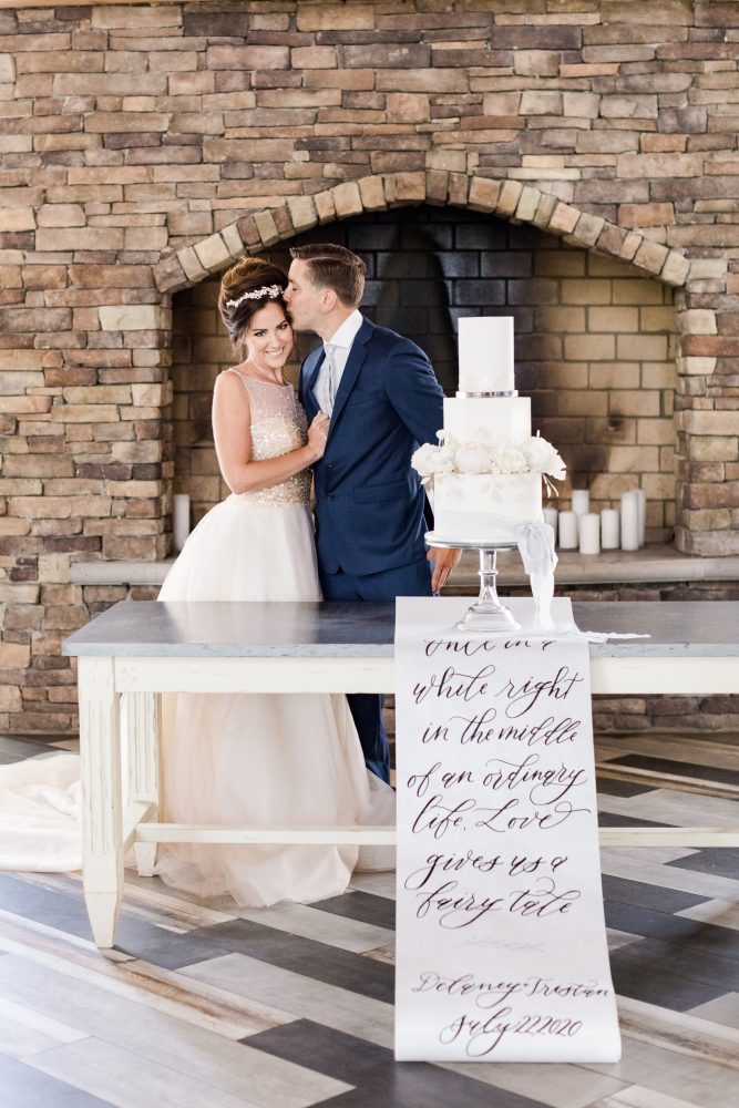 CoachHouseStyledShoot_BKP_4971-2-2 (1) Bride with textured bridal updo and pearl hairpiece beside wedding cake and calligraphy sign at New Jersey reception